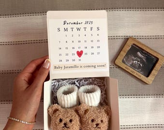Puede incluir: Una caja de regalo blanca que contiene dos patucos de bebé de peluche color canela con forma de oso y puños acanalados blancos. La tapa muestra un calendario de diciembre de 2025 con un corazón rojo en el día 24 y el texto "Baby Jaramillo is coming soon!".