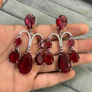 Peut inclure: Une paire de boucles d'oreilles pendantes en argent avec plusieurs pierres précieuses rouges en forme de goutte. Les boucles d'oreilles ont un design délicat et complexe avec de plus petites pierres précieuses entourant les plus grandes.