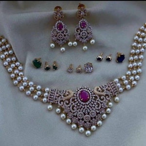 May include: A gold necklace with a large, ornate pendant featuring a pink stone surrounded by white stones. The necklace is made of three strands of pearls and includes a pair of matching earrings with pink and white stones.