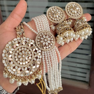 Könnte beinhalten: Eine weiße Perlenkette mit einem goldenen Anhänger mit einem Spiegeldesign. Die Kette wird mit passenden goldenen Ohrringen mit Spiegeldesign und Perlenakzenten kombiniert.