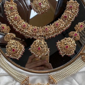 May include: A gold bridal jewelry set with pink and white stones. The set includes a necklace, earrings, a maang tikka, and a layered pearl choker.