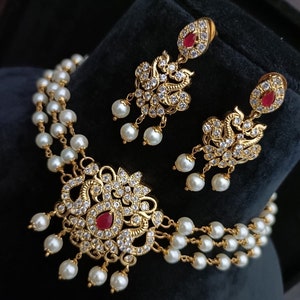 May include: A gold necklace with a red stone pendant and pearl accents. The necklace is layered with three strands of pearls. Matching earrings with red stones and pearls are also included.