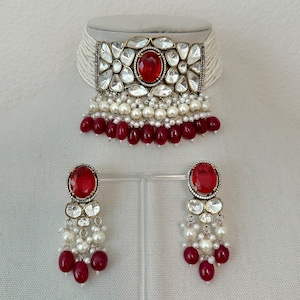 Op de afbeelding: Een witte parelketting met een rode edelsteen en heldere kristallen versieringen. Passende oorbellen met rode edelstenen en heldere kristallen hangen aan witte parels.