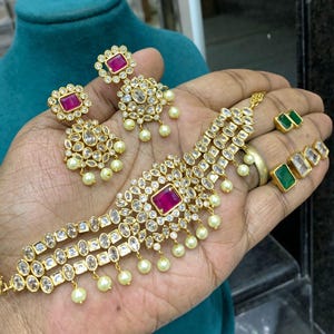 May include: A gold choker necklace with three rows of clear stones and a pink stone in the center. The necklace is accented with small white pearls. Matching earrings with pink stones and clear stones are also included.