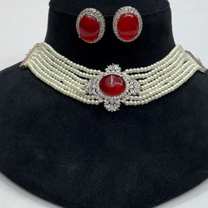 May include: A jewellery set comprising a multi-strand pearl necklace with a large, oval, red gemstone pendant and matching stud earrings. The pendant and earrings are set with clear crystals. The set is displayed on a black mannequin.