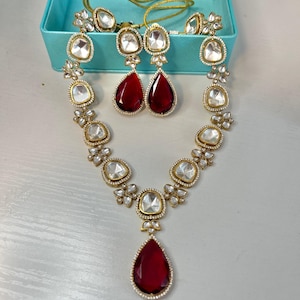 May include: A gold-toned jewelry set featuring a necklace, earrings, and rings. The necklace and earrings have large, teardrop-shaped, deep red gemstones. The necklace and earrings are accented with clear, square-cut stones and floral designs.