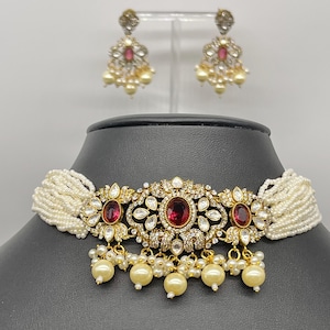 Peut inclure: Un collier ras de cou en perles blanches avec un pendentif en or et pierres rouges. Le pendentif présente un motif floral avec plusieurs pierres rouges. Le collier est accompagné d'une paire de boucles d'oreilles assorties avec des pierres rouges.
