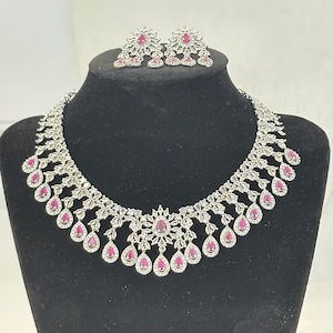 May include: A silver-toned necklace and matching earrings set. The necklace features a floral design with teardrop-shaped, ruby-colored stones and clear crystals. The earrings have a similar floral design.