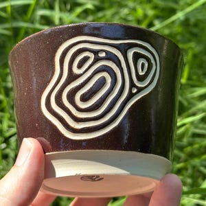 Może przedstawiać: Ciemnobrązowy ceramiczny kubek z błyszczącym wykończeniem, ozdobiony rzeźbionym, kremowym abstrakcyjnym wzorem topograficznym z boku. Podstawa kubka jest nieszkliwiona, odsłaniając jasną glinę, i zawiera mały wytłoczony znak na spodzie.