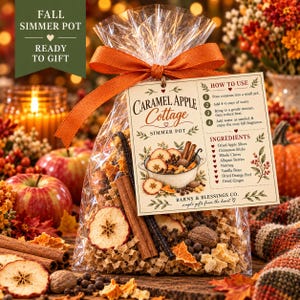 May include: A clear plastic bag containing a Caramel Apple Cottage simmer pot mix, tied with an orange ribbon. The attached tag lists ingredients including dried apple slices, cinnamon sticks, cloves, allspice, nutmeg, vanilla, and ginger.