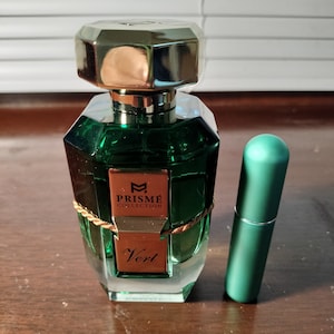 Puede incluir: Un frasco de perfume de vidrio verde con una tapa dorada y una etiqueta dorada que dice "Prisme Collection Vert". El frasco está sobre una superficie de madera junto a un tubo verde más pequeño.