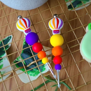Peut inclure: Deux pinces à cheveux en perles arc-en-ciel avec des breloques de montgolfières. Les perles sont violettes, rouges, orange, jaunes et vertes. Les montgolfières sont blanches avec des rayures orange, rouges et violettes.