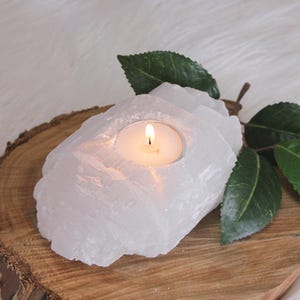 May include: A white, translucent, raw selenite crystal candle holder with a rough, jagged exterior texture. A small, round, lit tea light candle with a visible yellow flame sits in the centre cavity of the stone. The crystal has a fibrous appearance.