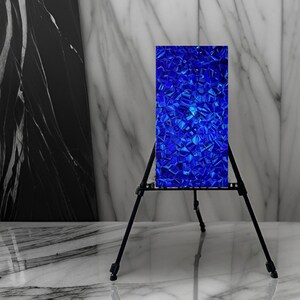 Op de afbeelding: Een blauwe abstracte kunstprint op een zwarte ezelstandaard. De print is gemaakt van veel kleine blauwe vierkanten.