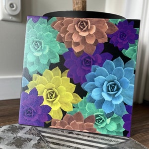 Pode incluir: Um azulejo quadrado colorido com um padrão de várias suculentas em tons de verde, roxo, amarelo e marrom. As suculentas estão dispostas em um padrão aleatório sobre um fundo preto.