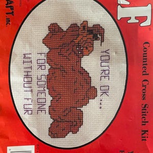 May include: A counted cross stitch kit featuring a brown bear with the text "You're OK... For someone without fur". The kit includes 14 count white Aida fabric, embroidery floss, a needle, and instructions. The kit is designed to fit a 4 inch by 5 1/2 inch oval or rectangle frame.