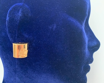 May include: A gold-coloured, rectangular stud earring with a geometric design. The earring is displayed on a blue velvet surface, showcasing its polished finish and design. The earring is a fashion accessory.