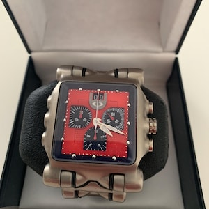 May include: A silver Oakley wristwatch with a square red face and black subdials. The watch has a black leather strap and is presented in a black box with a white interior. The watch face displays the number 09.