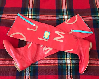 Puede incluir: Un par de botas rojas de caña alta con un logotipo blanco y un ribete azul. Las botas están hechas de cuero perforado.