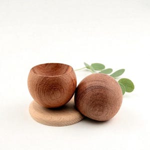 May include: Two small, round wooden bowls with a natural finish. One bowl is open, revealing the interior, while the other is a solid sphere. They rest on a light-colored wooden coaster, with green leaves in the background.