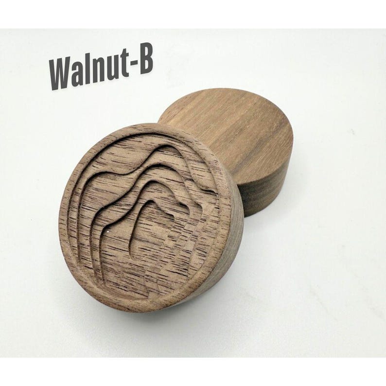 May include: Two wooden, circular objects. One has a carved mountain range design. The other is a plain, solid circle. The text "Walnut-B" is in the upper left corner. The objects are made of a light brown wood.