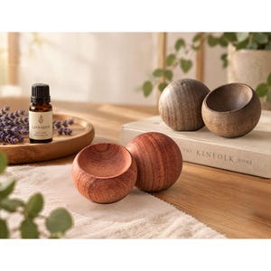 May include: Three spherical wooden bowls with concave centers, featuring natural wood grain patterns in varying shades of brown. One bowl is reddish-brown, while the other two are darker, muted brown. A small glass bottle of lavender essential oil is nearby.
