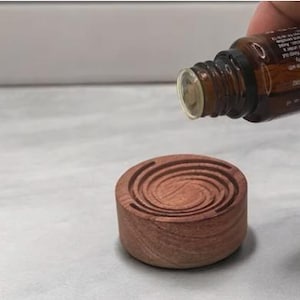 May include: A small, round, wooden essential oil diffuser with a carved spiral design. A brown glass bottle is held above the diffuser, with oil being poured into it. The diffuser is made of a reddish-brown wood.