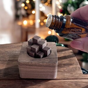 May include: A wooden cube puzzle with a small hole in the top. A hand is pouring essential oil into the hole. The puzzle is sitting on a wooden surface.