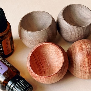 May include: Four small, wooden bowls in varying shades of brown and gray are arranged with two essential oil bottles. One bottle is labeled "Wild Orange" and the other "Lavender". The bowls have a smooth, rounded shape with a hollowed-out interior.