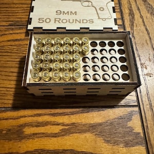 May include: A wooden box with a laser-cut design of a handgun and the text "9MM 50 ROUNDS." The box is open, revealing rows of brass-colored bullets. The box is sitting on a wooden surface.