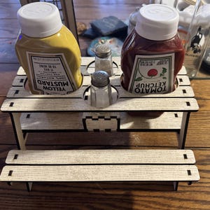 May include: A white wooden picnic table condiment holder with two glass salt shakers, a bottle of yellow mustard labeled "Yellow Mustard 100% Natural", and a bottle of red ketchup labeled "Tomato Ketchup".