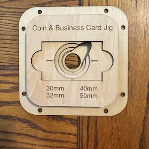 Puede incluir: Una plantilla de madera clara para monedas y tarjetas de visita, con un recorte circular central y un brazo giratorio. Incluye medidas para 30 mm, 32 mm, 40 mm y 50 mm. La plantilla tiene esquinas redondeadas y pequeños agujeros.