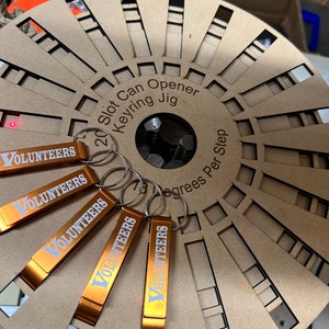 May include: Five orange metal bottle openers with the word "VOLUNTEERS" printed in white on each. The openers are attached to key rings and are hanging on a wooden jig with the text "20 Slot Can Opener Keyring Jig 78 Degrees Per Step".
