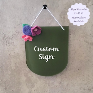 May include: A green custom sign with rounded edges, measuring 6 inches by 4.75 inches. The sign is decorated with felt flowers in pink, purple, and green. The sign reads "Custom Sign" in white lettering.