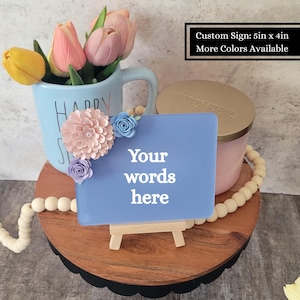 May include: A rectangular blue custom sign measuring 5 inches by 4 inches, featuring white text that reads 'Your words here'. The sign is decorated with three paper flowers in shades of pink, light blue, and lavender on the top left corner.