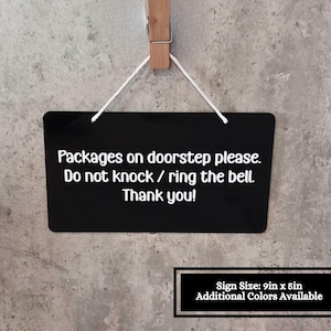 May include: A rectangular black sign with white text that reads: Packages on doorstep please. Do not knock / ring the bell. Thank you! The sign is suspended by a white string from a wooden clip and measures 9 inches by 5 inches in size.