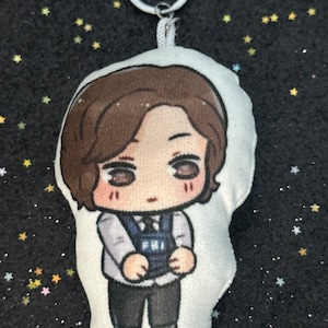May include: A plush keychain featuring a cartoon character with brown hair, wearing a white shirt, dark vest, and black pants. The keychain has a silver metal ring and a white fabric loop for attachment. The background is black with scattered star-shaped glitter.