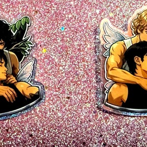May include: Two stickers featuring illustrations of male figures with angel wings, embracing. One has dark hair, the other blonde. They are set against a glittery pink background. The figures are in a romantic embrace, with one figure resting his head on the other's shoulder.