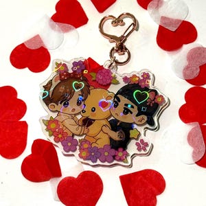 May include: A clear acrylic keychain featuring a holographic illustration of two chibi characters hugging a yellow teddy bear, surrounded by colorful flowers. The keychain includes a rose gold heart-shaped clasp at the top for attachment.