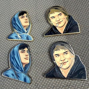 May include: Four enamel pins featuring a portrait of a person. Two pins show the person in a blue hoodie, and two show the person in a beanie and a black hoodie with a cigarette. All pins have a gold border.