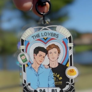 May include: A clear acrylic keychain featuring two men in a heart-shaped design with the text "THE LOVERS". The keychain has a black clasp and includes the numbers 24 and 81. The design is set against a blue and white background.