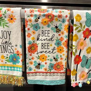 May include: Three white kitchen towels with floral and bee designs. One towel reads "ENJOY the little THINGS" with a fringe. Another says "BEE kind BEE sweet". The third features butterflies.