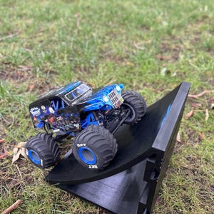 May include: A blue and black monster truck toy with large tires is positioned on a black ramp. The truck has a detailed design with blue flames and graphics. The tires have the text "BKT" on them. The scene is set outdoors on grass.