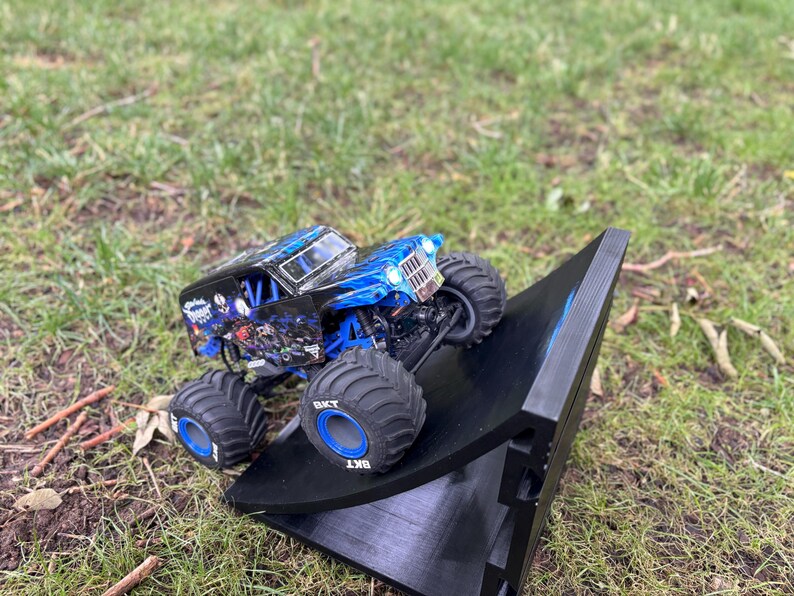 May include: A blue and black monster truck toy with large tires is positioned on a black ramp. The truck has a detailed design with blue flames and graphics. The tires have the text "BKT" on them. The scene is set outdoors on grass.