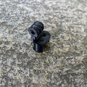 May include: A small, black, 3D-printed object with a cylindrical base, a circular top, and a connecting piece. The object appears to be a specialized component or fitting, possibly for a larger assembly. The surface has a textured appearance.