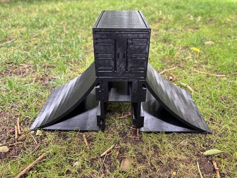 May include: A black, 3D-printed ramp structure with a rectangular top and angled side panels. The top section has a textured surface, and the side panels curve outward. The structure is designed to be placed on the ground.
