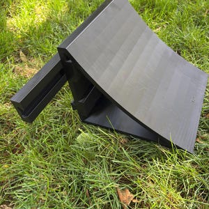 May include: Two black, wedge-shaped wheel chocks rest on green grass. The chocks are made of a solid material and have a ridged surface. One is angled, and the other is flat. The chocks are designed to prevent vehicle movement.