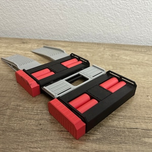 May include: A 3D-printed device in black, red, and grey. It has two rectangular sections with red cylinders, linked by a grey bridge. The ends are red and angled, with a textured surface.