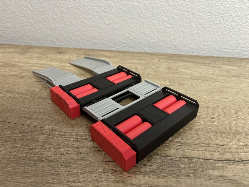 May include: A 3D-printed device with a black, red, and gray color scheme. The design features two rectangular sections with red cylinders, connected by a gray bridge. The ends are red and angled.