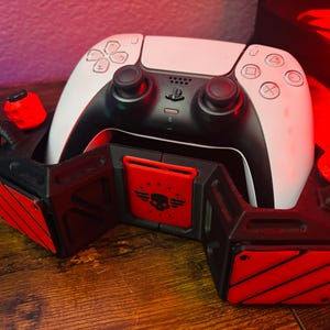 Puede incluir: Un mando de PlayStation 5 DualSense blanco está montado en un soporte de impresión 3D negro y rojo. El soporte tiene un logotipo de calavera en la parte delantera y detalles rojos.